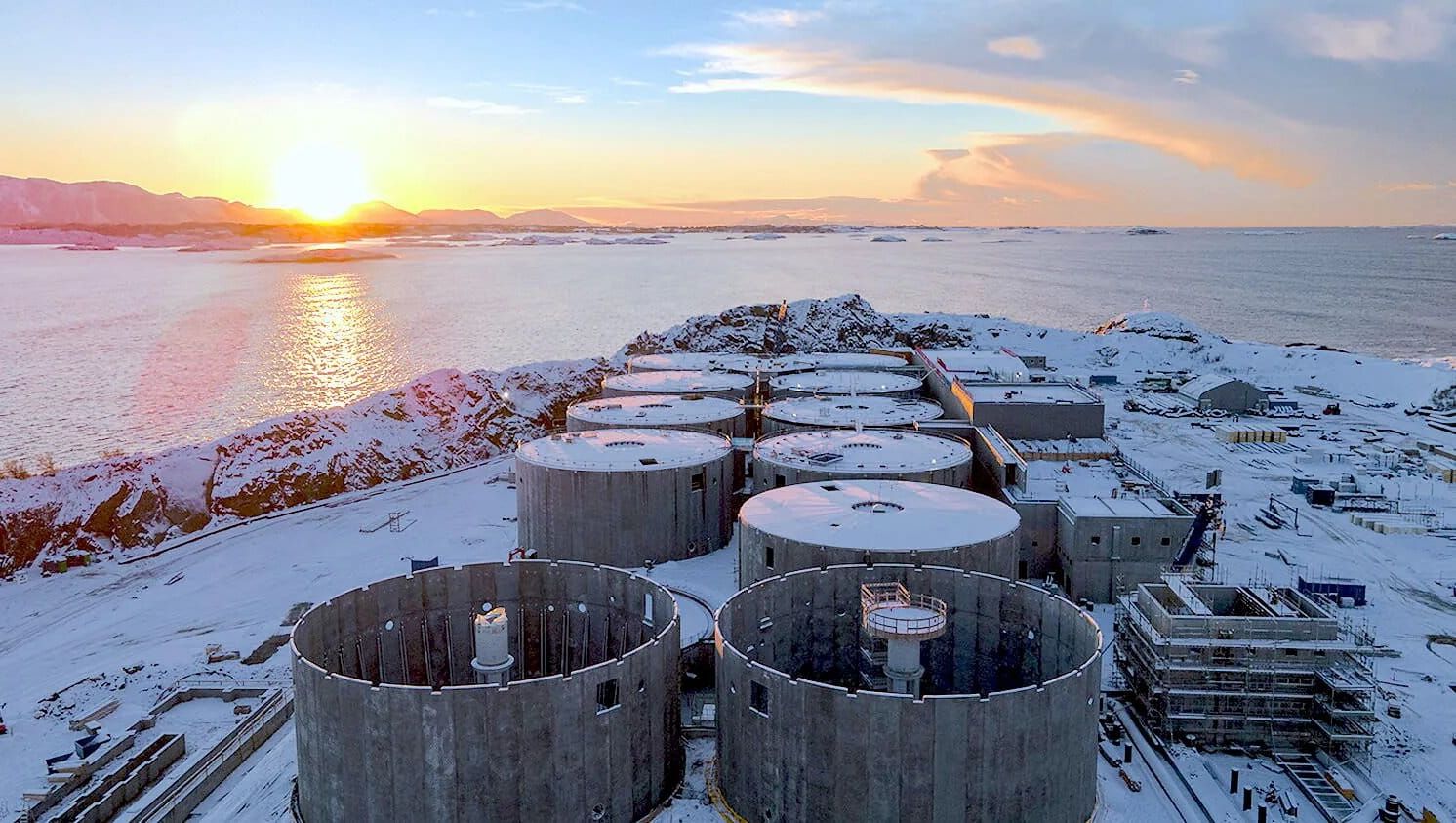Snødekt fiskeoppdrettsanlegg på land med solnedgang over sjø